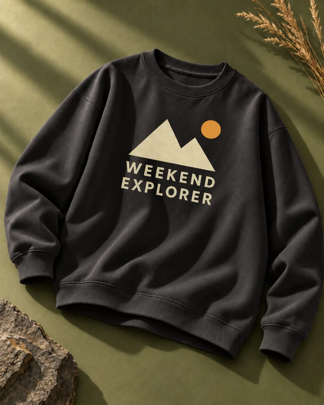 苔绿色与石板背景中的黑色圆领卫衣，印有山峰太阳与WEEKEND EXPLORER